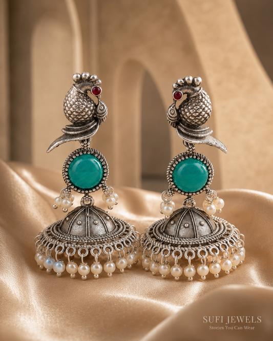 The Bird Carried Bells silver-look jhumka earrings with a sculpted bird motif, teal centre stone and white pearl detailing.